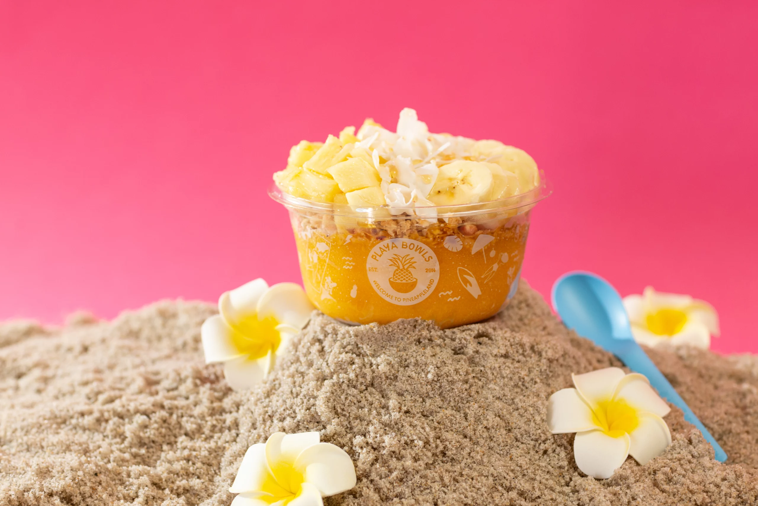 Mango bowl with banana, pineapple, and coconut on a small pile of sand with a blue spoon